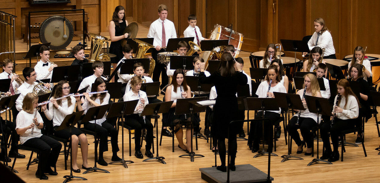 Symphonic Band performing onstage during a concert. Their conductor stands in front with her arms raised.