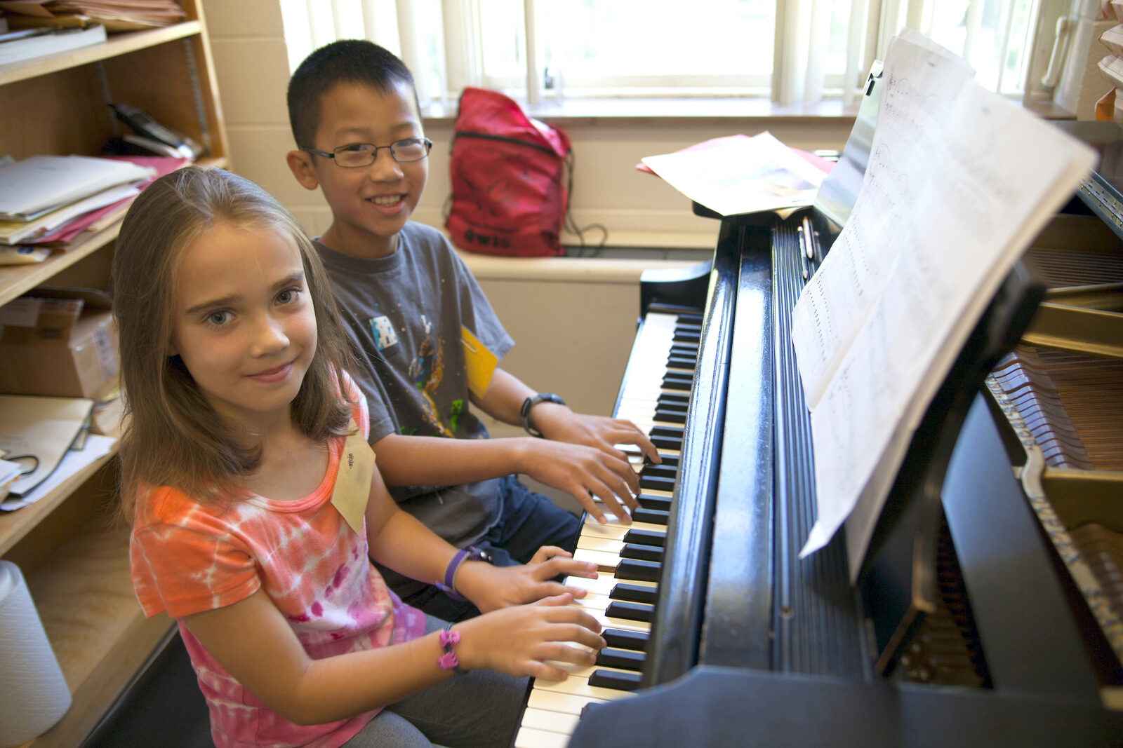 A young boy and girl sit at a piano in a studio. Both have their hands on the keys and smile at the camera.