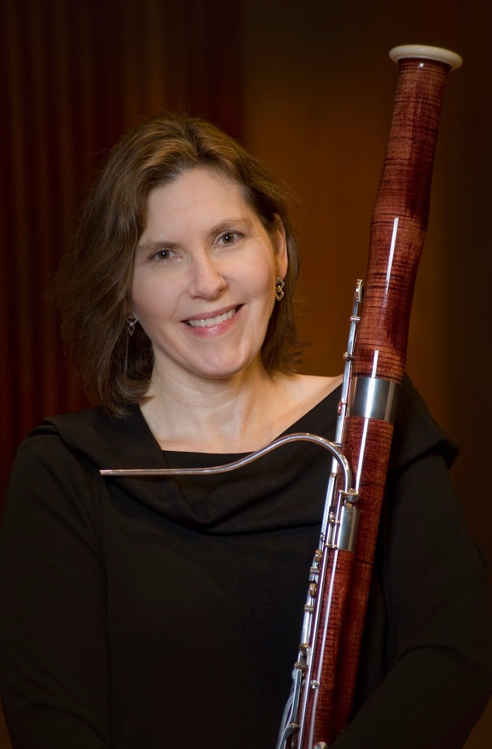 A woman with shoulder-length, wavy hair holds a bassoon and smiles at the camera.