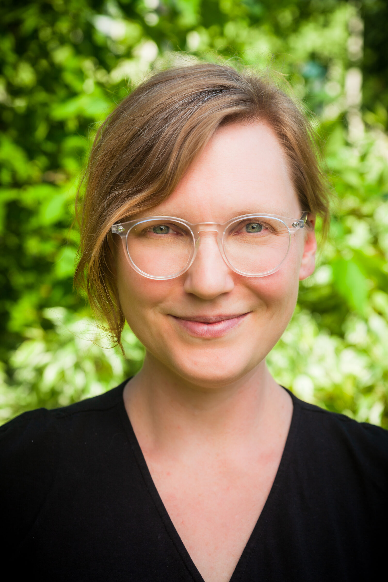 A headshot of a woman with short hair and glasses. She stands outside and smiles at the camera.
