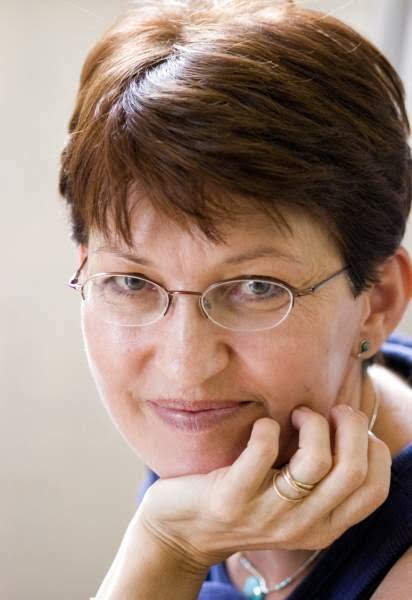 A woman with short, brown hair and glasses smiles at the camera and poses leaning with her chin in her hand.