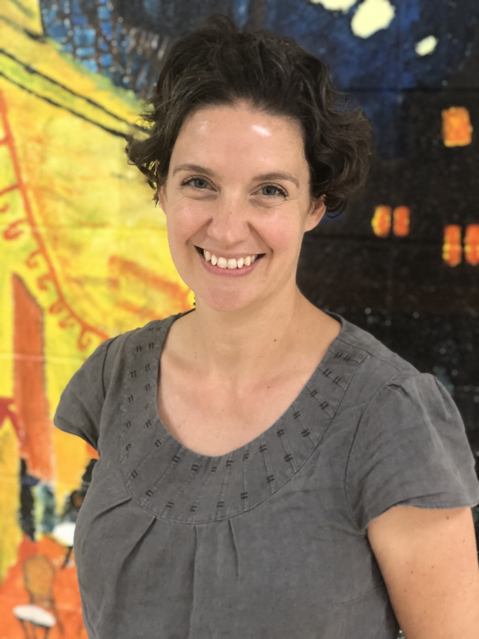 A headshot of a smiling woman with dark, wavy hair standing in front of a colorful mural.