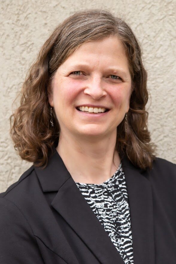 A smiling woman with wavy, brunette hair wears a black blazer.
