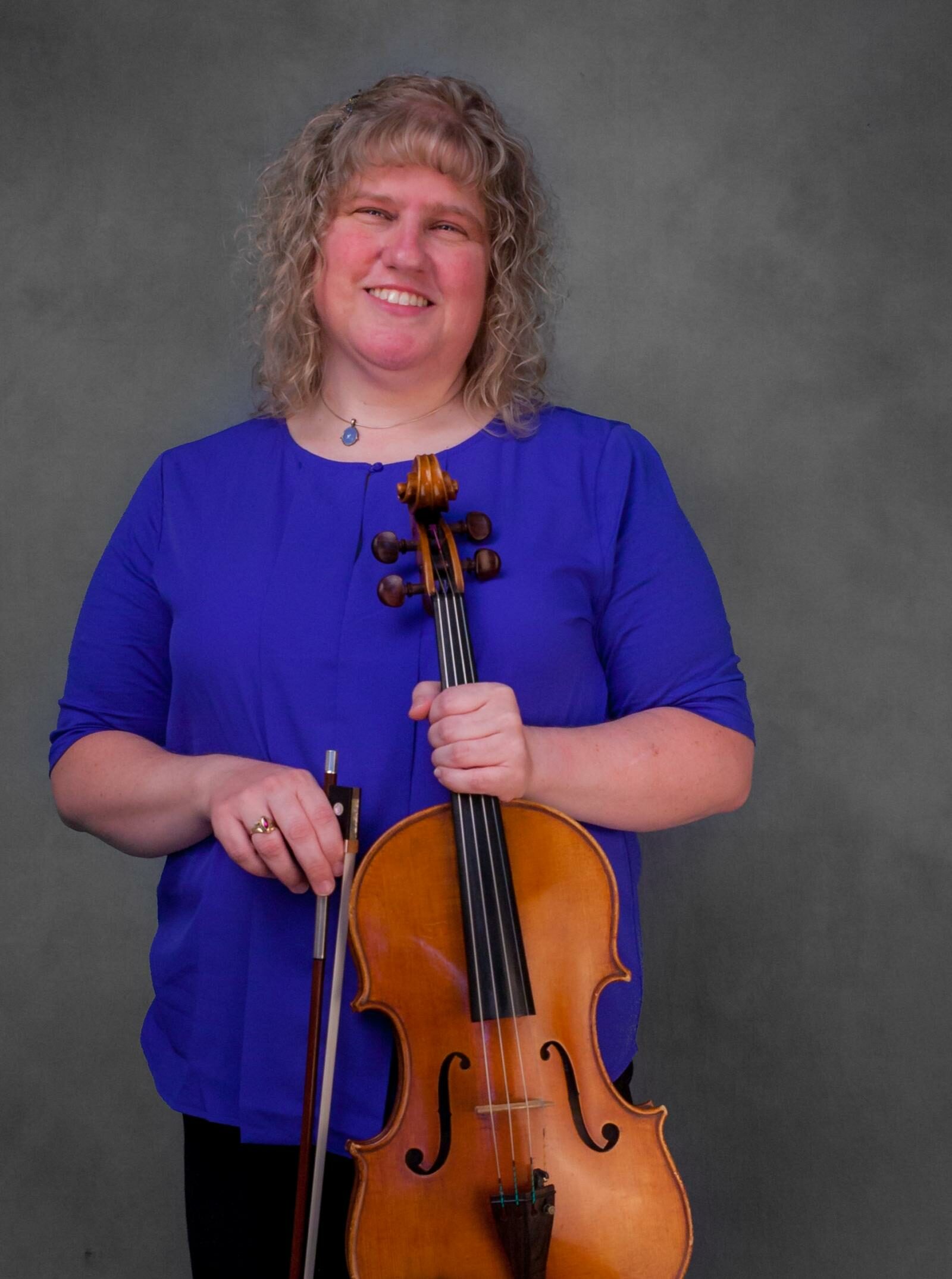 A smiling woman with curly hair and a blue dress shirt holds a viola. 