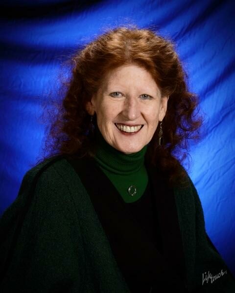 A headshot of a smiling woman with curly red hair. She wears a green turtleneck and sits in front of a blue backdrop.