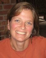 A headshot of a smiling woman in a burnt orange sweater. She's sits in front of a brick background.