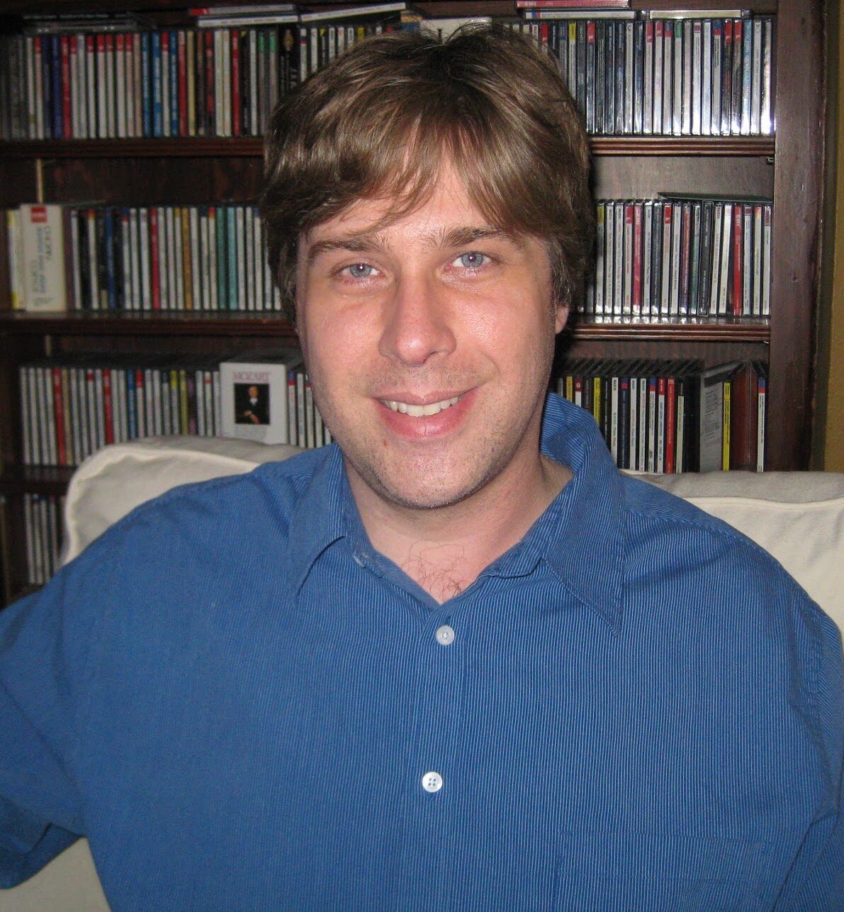A man in a blue shirt smiles at the camera while sitting in a chair.
