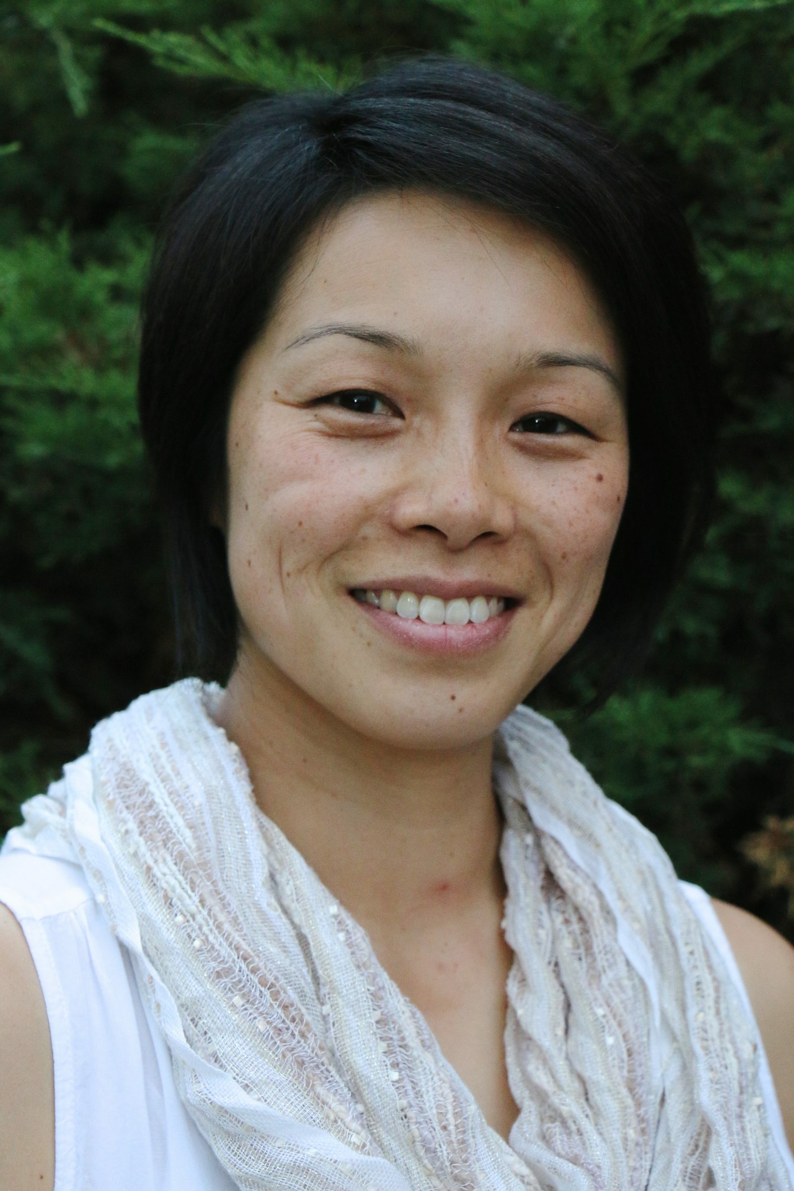 A photo of a smiling woman with short black hair. She wears a scarf and stands in front of a backdrop of green foliage.