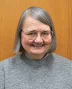 A photo of a smiling woman with short, gray hair and glasses. She's wearing a grey sweatshirt.