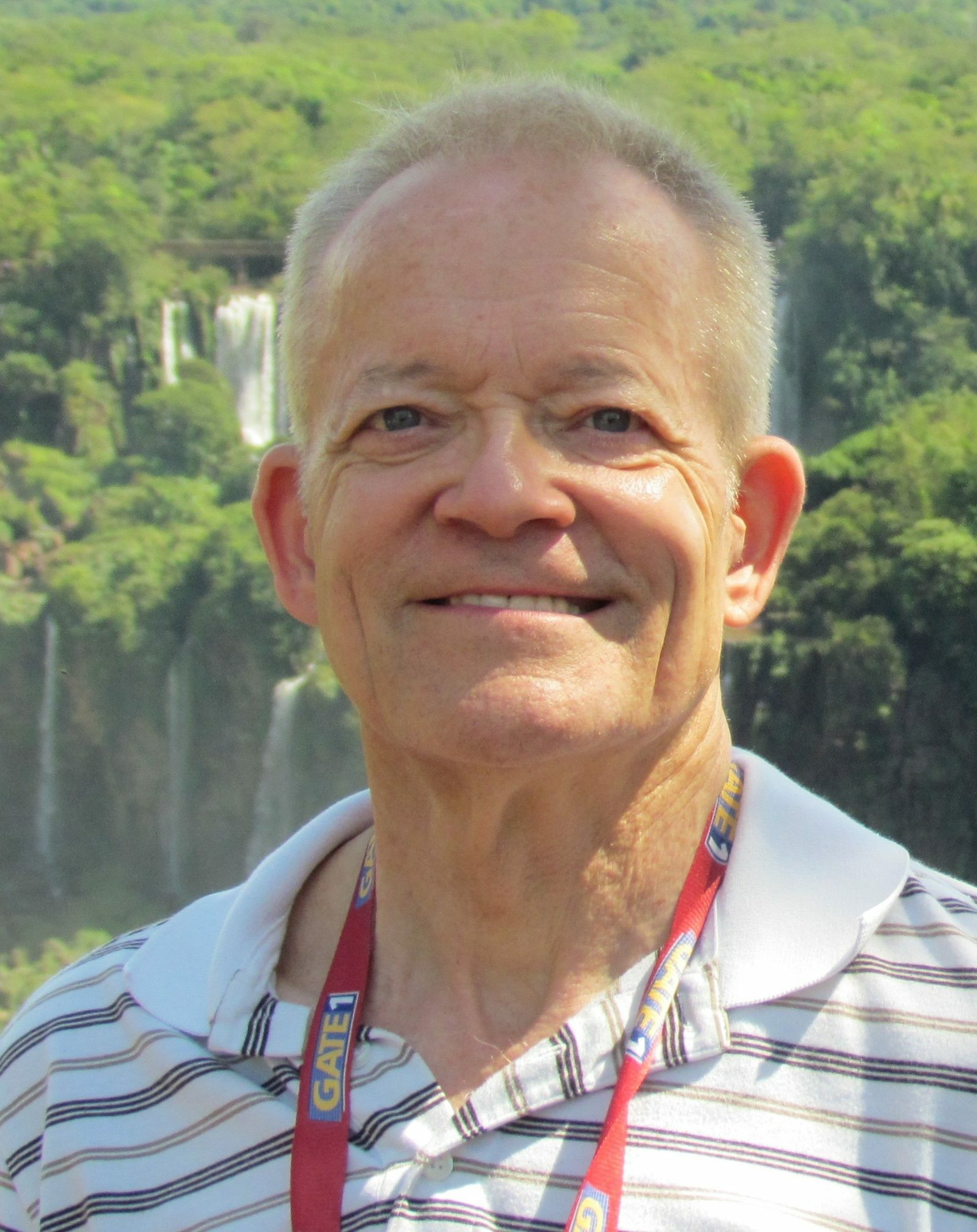 A headshot of a man with short, gray-blonde hair. He wears a striped shirt and stands outside in front of lush, green trees.