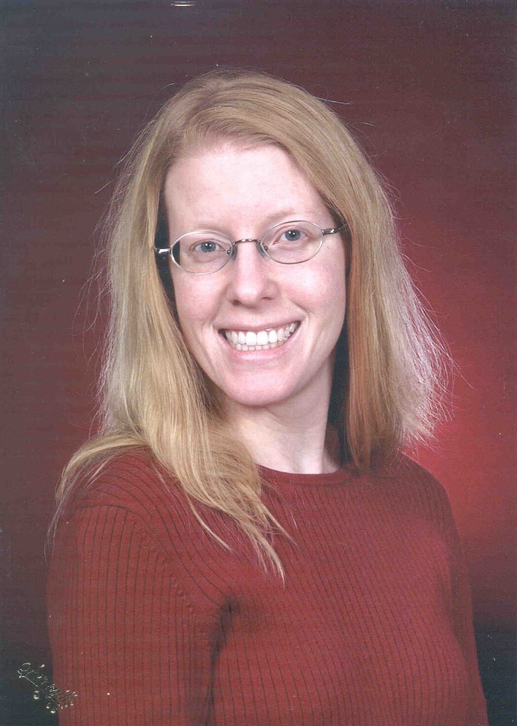 A blonde woman with glasses smiles at the camera in front of a soft red backdrop.