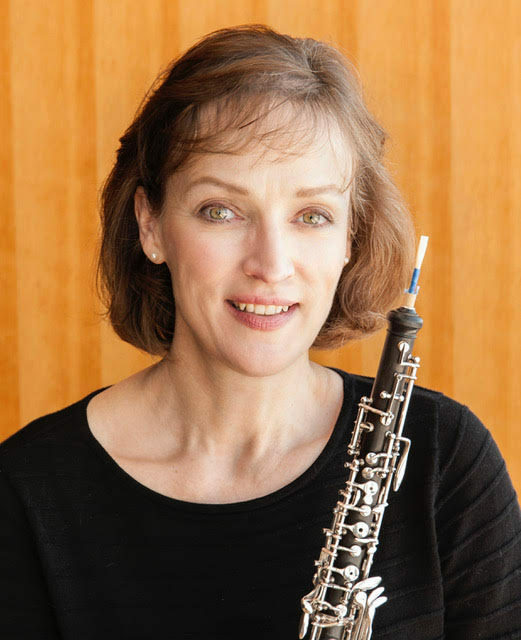 A headshot of a smiling woman with wavy brown hair. She holds an oboe and stands in front of a woodgrain backdrop.