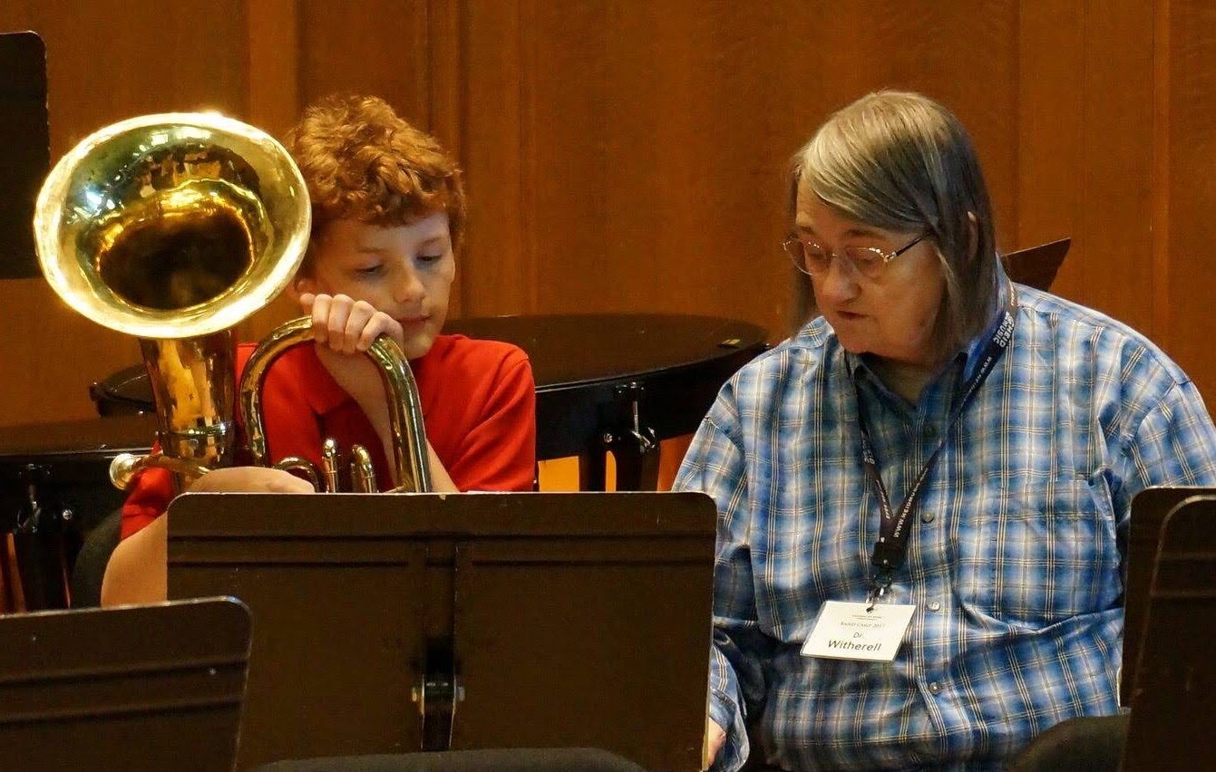 A student with a tuba and his instructor sit behind a music stand. His instructor speaks and points at the score.