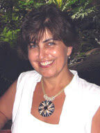 A woman with short brown hair smiles at the camera standing in front of a lush green foliage.
